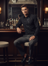 Man in a bar setting wearing a black sequined shirt and gray pants.
