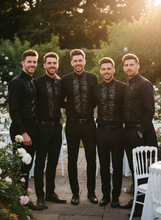Five men in matching black outfits standing together outdoors with greenery and flowers in the background.
