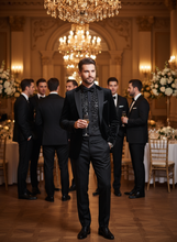 Man in a black tuxedo standing in an elegant room with chandeliers and other formally dressed individuals.