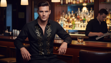 Man in a stylish black jacket with gold embroidery sitting at a bar.